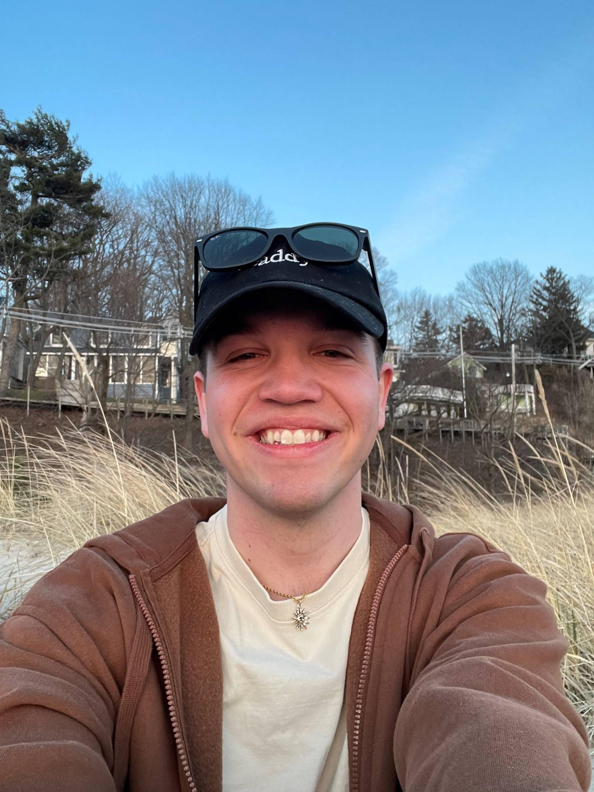 young man with black baseball hat and sunglasses smiling with houses in the background